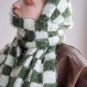 May include: A hooded scarf featuring a checkered pattern in forest green and white. The accessory is crafted from a combination of smooth yarn and fuzzy, textured yarn, creating a distinct contrast in the square blocks throughout the design.