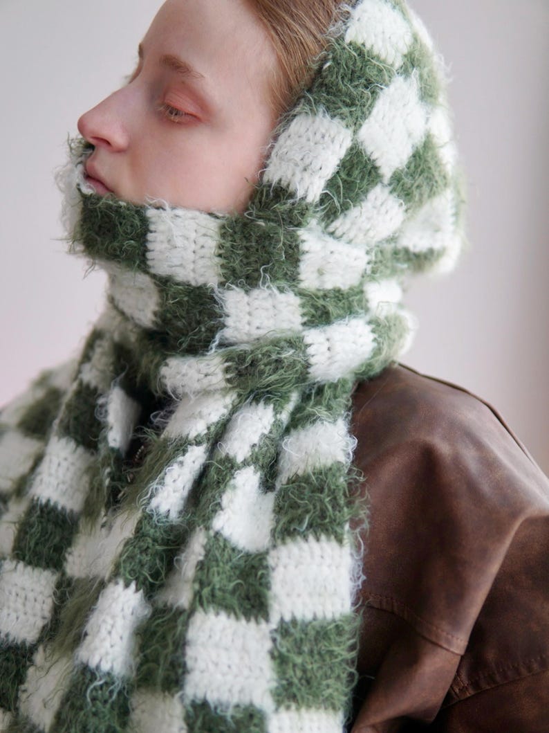 May include: A hooded scarf featuring a checkered pattern in forest green and white. The accessory is crafted from a combination of smooth yarn and fuzzy, textured yarn, creating a distinct contrast in the square blocks throughout the design.