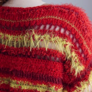 May include: A crochet sweater featuring horizontal stripes in vibrant red, deep burgundy, and bright lime green. The garment is constructed with a mix of textured, fuzzy yarn and open-work stitch patterns, creating a layered, multi-dimensional appearance.