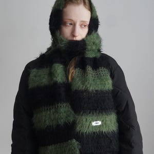 May include: A fuzzy, hooded scarf featuring horizontal stripes in alternating black and olive green. The knit texture is soft and voluminous, with a small white rectangular label stitched onto the lower section displaying the text ZARA.