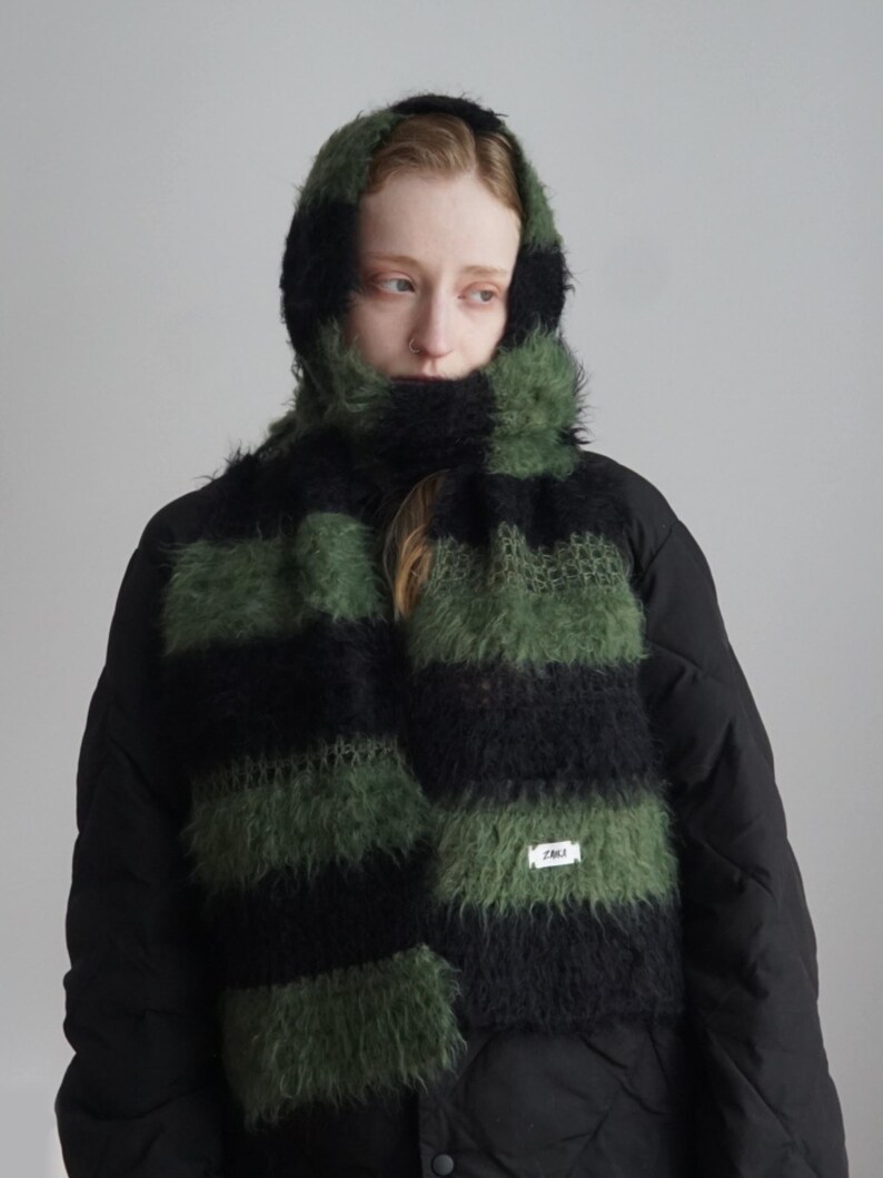 May include: A fuzzy, hooded scarf featuring horizontal stripes in alternating black and olive green. The knit texture is soft and voluminous, with a small white rectangular label stitched onto the lower section displaying the text ZARA.