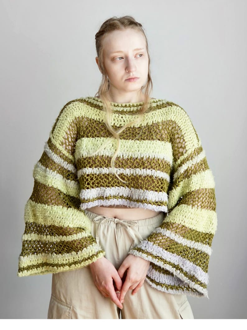 May include: A cropped, long-sleeved crochet jumper featuring horizontal stripes in shades of olive green, pale yellow, and light lavender. The garment has a loose, open-knit texture with wide, flared sleeves and a wide, rounded neckline.