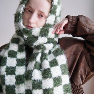 May include: A hooded scarf featuring a checkered pattern in forest green and white. The accessory is crafted from a combination of smooth yarn and a fuzzy, textured yarn, creating a distinct contrast in the square blocks throughout the design.