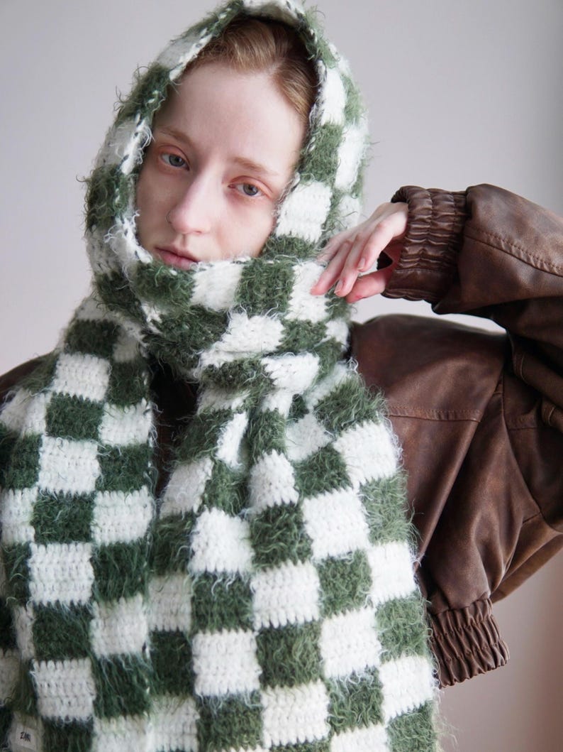 May include: A hooded scarf featuring a checkered pattern in forest green and white. The accessory is crafted from a combination of smooth yarn and a fuzzy, textured yarn, creating a distinct contrast in the square blocks throughout the design.