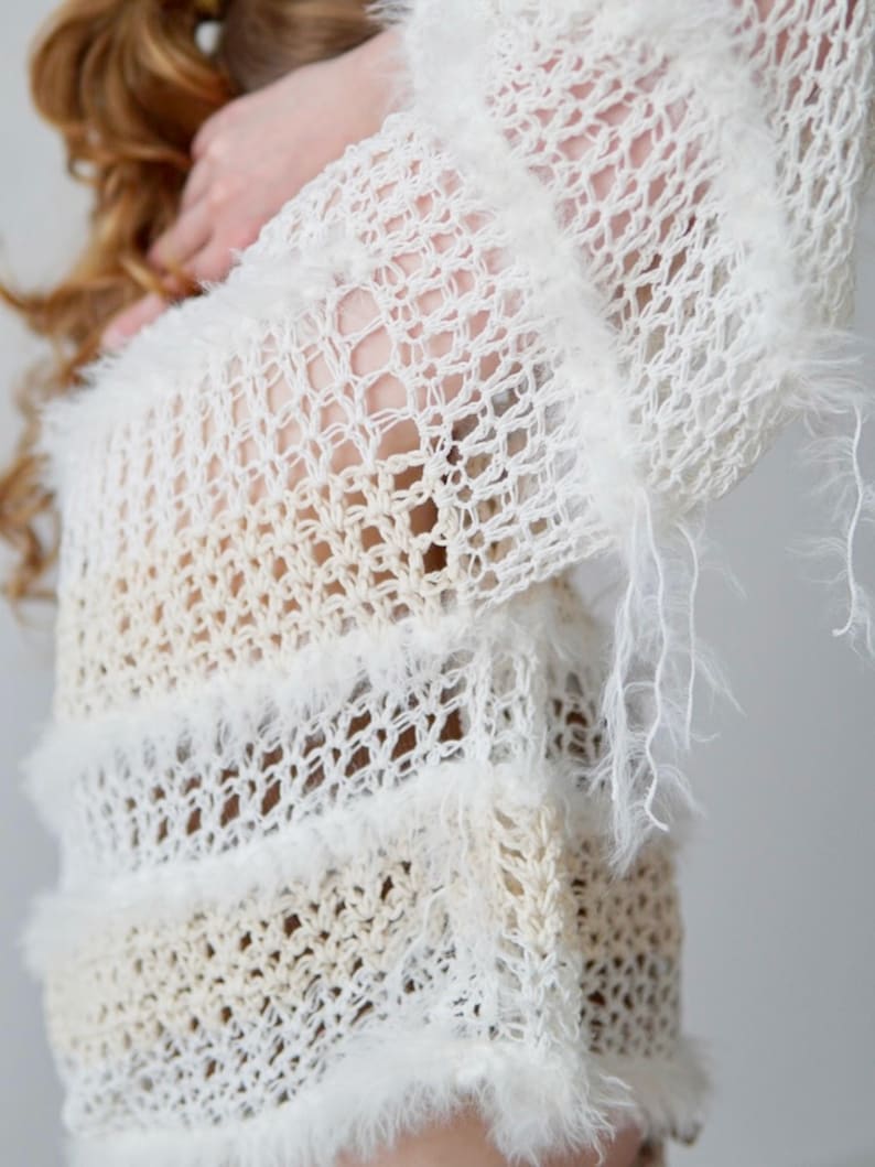 May include: A long-sleeved, open-knit jumper in cream and off-white tones. The garment features a loose, airy crochet-style weave with horizontal bands of fuzzy, textured yarn trim that add dimension to the lightweight, semi-sheer fabric.