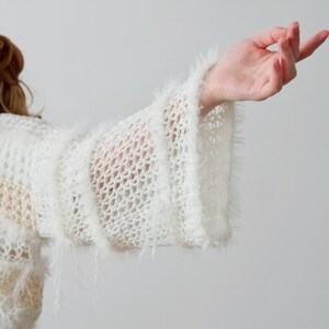 May include: A white, open-knit jumper featuring wide, flared sleeves with a fuzzy, textured trim. The garment is constructed with a loose, net-like pattern and includes horizontal bands of soft, shaggy yarn detailing along the sleeves.