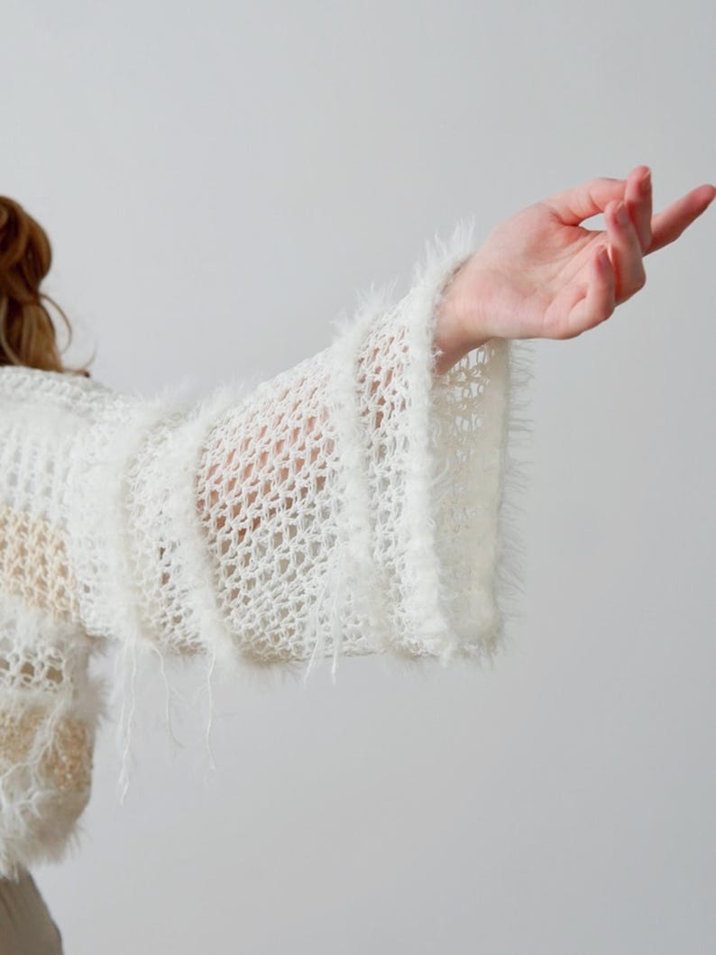 May include: A white, open-knit jumper featuring wide, flared sleeves with a fuzzy, textured trim. The garment is constructed with a loose, net-like pattern and includes horizontal bands of soft, shaggy yarn detailing along the sleeves.