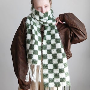 May include: A fuzzy, knitted scarf featuring a green and white checkered pattern. The scarf has a soft, textured appearance with white fringe tassels on one end and green fringe tassels on the other, and a small white rectangular label at the bottom.