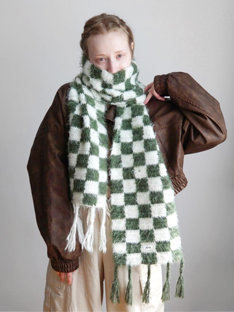 May include: A fuzzy, knitted scarf featuring a green and white checkered pattern. The scarf has a soft, textured appearance with white fringe tassels on one end and green fringe tassels on the other, and a small white rectangular label at the bottom.