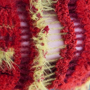 May include: A knitted textile featuring a textured, fuzzy yarn in vibrant red and bright yellow. The construction alternates between dense, shaggy sections of red and yellow fibers and open, ladder-like gaps that reveal the surface underneath.