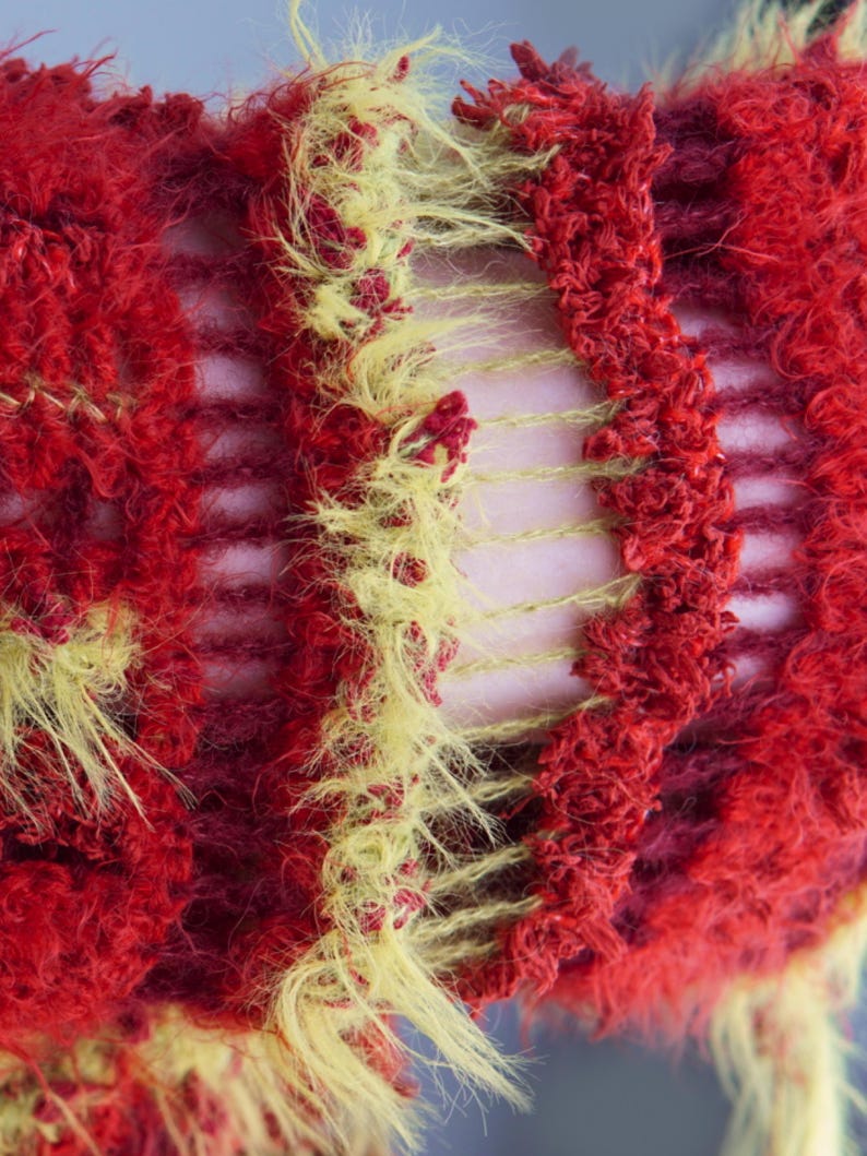 May include: A knitted textile featuring a textured, fuzzy yarn in vibrant red and bright yellow. The construction alternates between dense, shaggy sections of red and yellow fibers and open, ladder-like gaps that reveal the surface underneath.