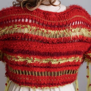 May include: A cropped, short-sleeved crochet top featuring horizontal stripes in shades of red and lime green. The garment displays a mix of textured, fuzzy yarn and open-work stitch patterns, with dangling lime green fringe accents at the sleeves.