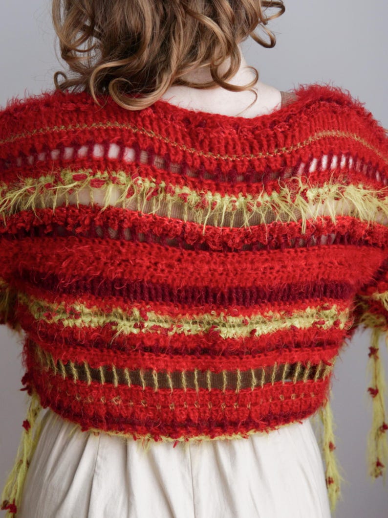 May include: A cropped, short-sleeved crochet top featuring horizontal stripes in shades of red and lime green. The garment displays a mix of textured, fuzzy yarn and open-work stitch patterns, with dangling lime green fringe accents at the sleeves.