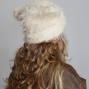 May include: A cream-colored beanie with a fuzzy, shaggy texture. The hat features a distinct knit construction with long, soft fibers that create a voluminous appearance. The beanie has a folded brim and a rounded top, providing a textured look.