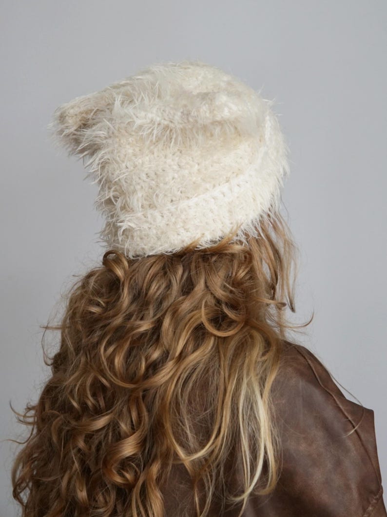 May include: A cream-colored beanie with a fuzzy, shaggy texture. The hat features a distinct knit construction with long, soft fibers that create a voluminous appearance. The beanie has a folded brim and a rounded top, providing a textured look.