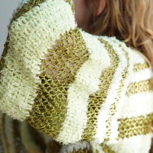 May include: A long-sleeved jumper featuring horizontal stripes in alternating textures and colours. The design includes thick, fuzzy bands in pale yellow and olive green, interspersed with sections of open, loose-knit olive green yarn.