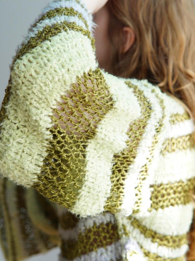 May include: A long-sleeved jumper featuring horizontal stripes in alternating textures and colours. The design includes thick, fuzzy bands in pale yellow and olive green, interspersed with sections of open, loose-knit olive green yarn.
