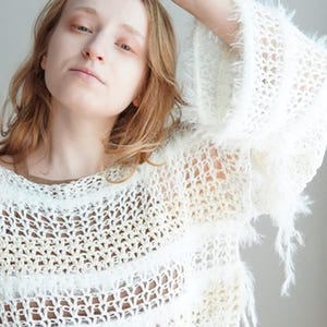 May include: A white long-sleeved jumper featuring an open-knit crochet pattern with alternating horizontal bands of fuzzy, textured yarn. The garment has a loose, relaxed fit with wide sleeves and a round neckline, showcasing a semi-sheer construction.