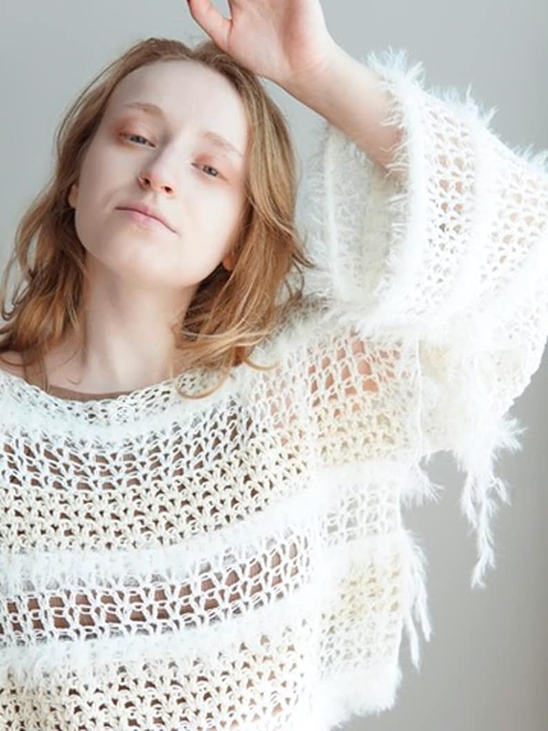 May include: A white long-sleeved jumper featuring an open-knit crochet pattern with alternating horizontal bands of fuzzy, textured yarn. The garment has a loose, relaxed fit with wide sleeves and a round neckline, showcasing a semi-sheer construction.