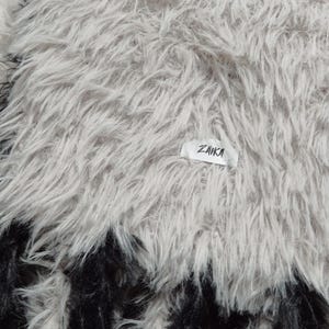 May include: A plush, long-pile faux fur throw blanket featuring a light gray and white color palette with a distinct black border at the bottom edge. A small white rectangular fabric tag with the black text ZAIKA is sewn onto the surface.