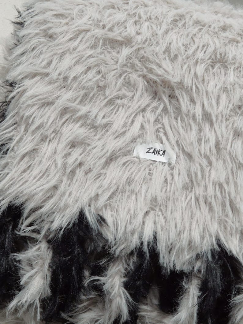 May include: A plush, long-pile faux fur throw blanket featuring a light gray and white color palette with a distinct black border at the bottom edge. A small white rectangular fabric tag with the black text ZAIKA is sewn onto the surface.