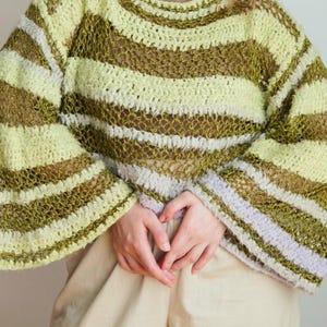 May include: A long-sleeve crochet jumper featuring horizontal stripes in shades of lime green, olive green, and light lavender. The garment has a loose, open-knit texture with wide sleeves and a wide neckline, layered over a solid tan shirt.