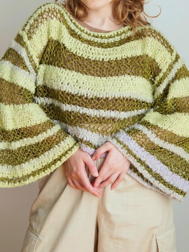 May include: A long-sleeve crochet jumper featuring horizontal stripes in shades of lime green, olive green, and light lavender. The garment has a loose, open-knit texture with wide sleeves and a wide neckline, layered over a solid tan shirt.
