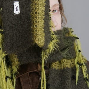 Op de afbeelding: Een donker olijfgroene en limoengroene gebreide sjaal met een pluizige, getextureerde garenconstructie en lange, losse franjes. Een wit rechthoekig stoffen label met de zwarte tekst ZAIKA is op het oppervlak van de sjaal genaaid.