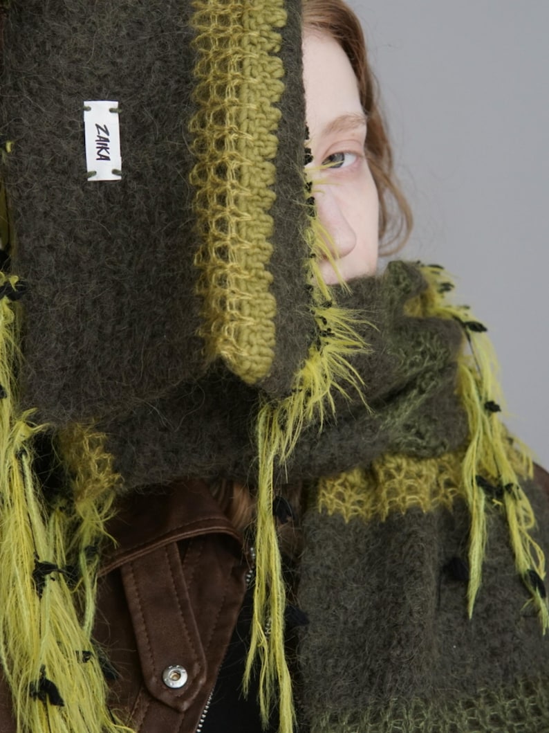 Op de afbeelding: Een donker olijfgroene en limoengroene gebreide sjaal met een pluizige, getextureerde garenconstructie en lange, losse franjes. Een wit rechthoekig stoffen label met de zwarte tekst ZAIKA is op het oppervlak van de sjaal genaaid.