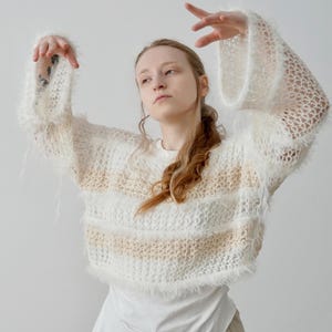 May include: A cropped, long-sleeved jumper featuring a loose, open-knit texture with alternating horizontal stripes of white fuzzy yarn and a light beige, finer yarn. The garment has wide, flared sleeves and a raw, shaggy hemline finish.