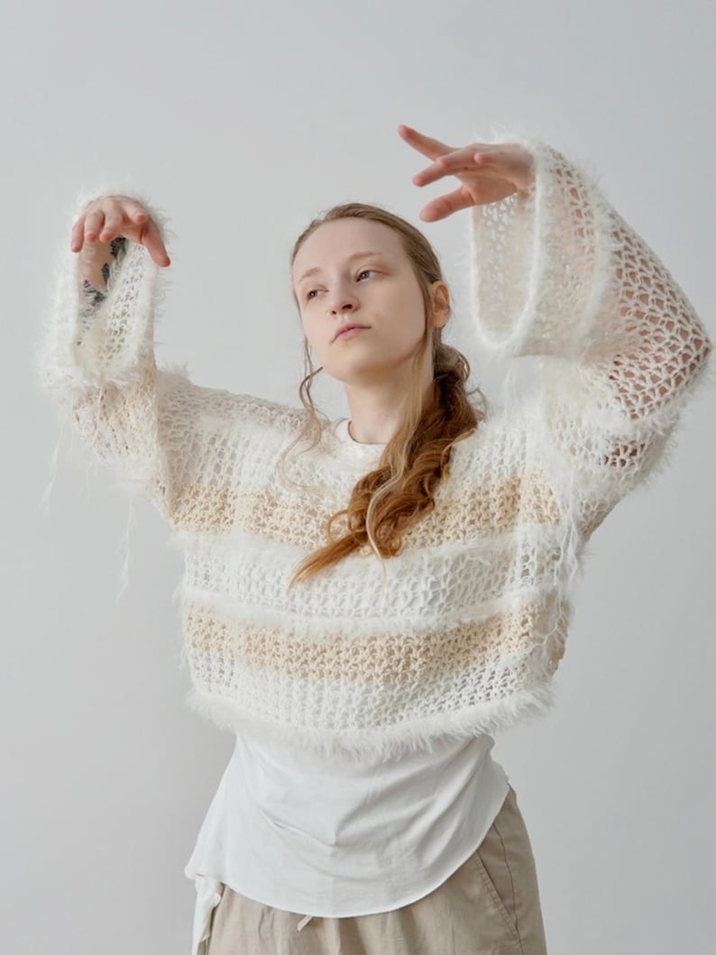 May include: A cropped, long-sleeved jumper featuring a loose, open-knit texture with alternating horizontal stripes of white fuzzy yarn and a light beige, finer yarn. The garment has wide, flared sleeves and a raw, shaggy hemline finish.