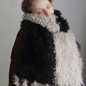 May include: A long, rectangular scarf made of thick, shaggy faux fur featuring a color-blocked design of alternating black and light beige sections. The ends of the scarf are finished with long, dangling fringe tassels in matching black and beige.