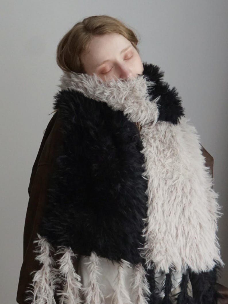 May include: A long, rectangular scarf made of thick, shaggy faux fur featuring a color-blocked design of alternating black and light beige sections. The ends of the scarf are finished with long, dangling fringe tassels in matching black and beige.