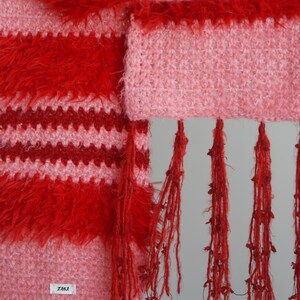 May include: A crochet scarf featuring alternating horizontal stripes of fuzzy red yarn and textured light pink yarn. The scarf includes long, dangling red fringe tassels along the bottom edge and a small white rectangular tag labelled ZAIKA.