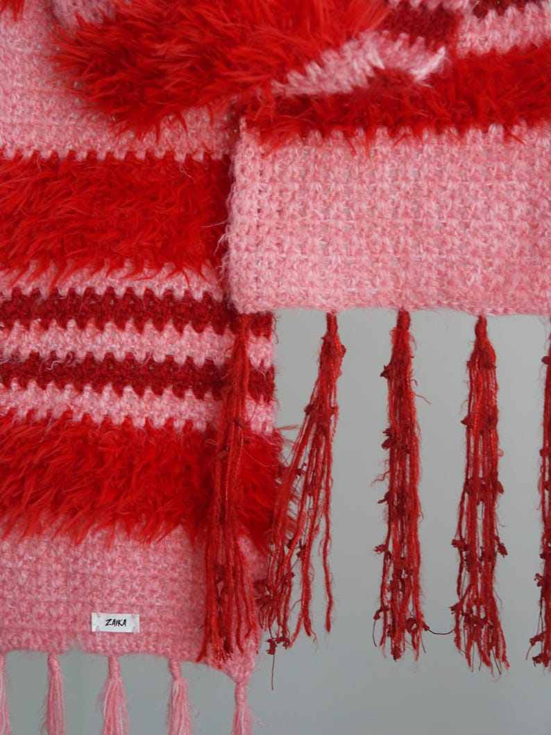 May include: A crochet scarf featuring alternating horizontal stripes of fuzzy red yarn and textured light pink yarn. The scarf includes long, dangling red fringe tassels along the bottom edge and a small white rectangular tag labelled ZAIKA.
