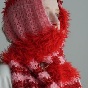 May include: A hooded scarf featuring a crochet pattern with alternating stripes of light pink and dark red yarn. The edges of the hood and the scarf are trimmed with a vibrant red faux fur texture, creating a soft and fuzzy appearance throughout.