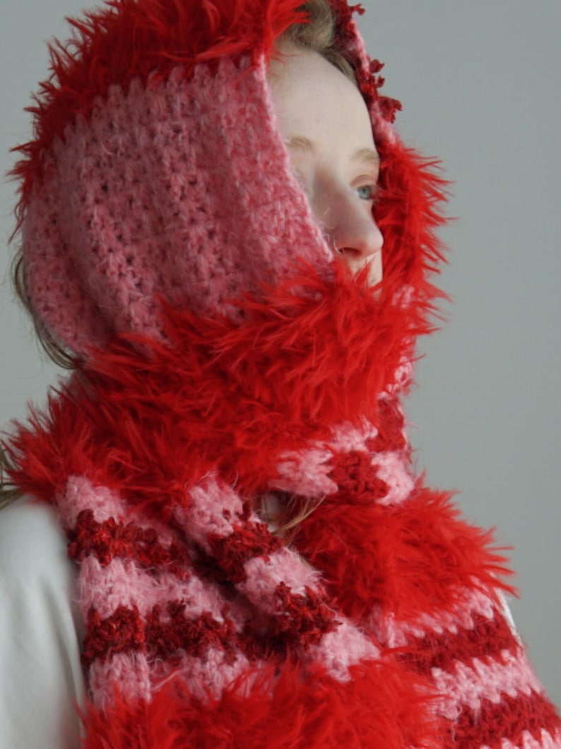 May include: A hooded scarf featuring a crochet pattern with alternating stripes of light pink and dark red yarn. The edges of the hood and the scarf are trimmed with a vibrant red faux fur texture, creating a soft and fuzzy appearance throughout.