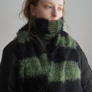 May include: A fuzzy, knitted scarf featuring wide, alternating horizontal stripes in black and olive green. The texture of the yarn is long and brushed, creating a soft, voluminous appearance that wraps around the neck over a black quilted jacket.