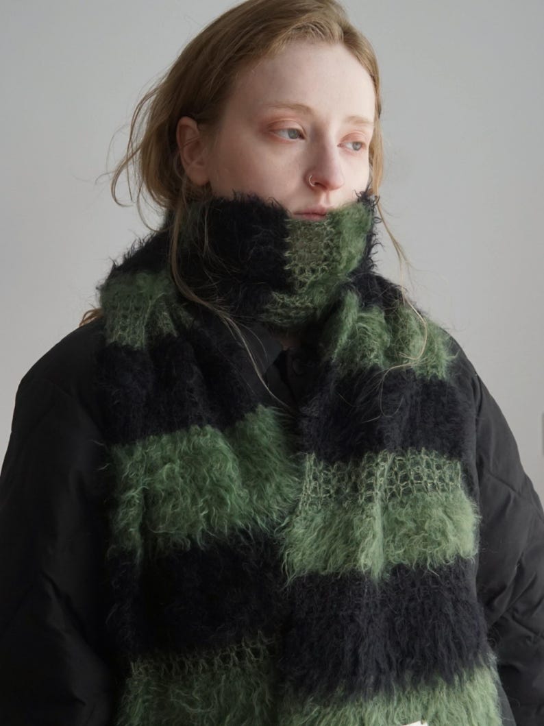 May include: A fuzzy, knitted scarf featuring wide, alternating horizontal stripes in black and olive green. The texture of the yarn is long and brushed, creating a soft, voluminous appearance that wraps around the neck over a black quilted jacket.