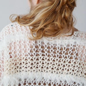 May include: A loose-knit crochet top featuring alternating horizontal bands of white and beige yarn. The garment displays a mix of open-work stitch patterns and textured, fuzzy yarn sections, creating a layered, semi-sheer appearance throughout.