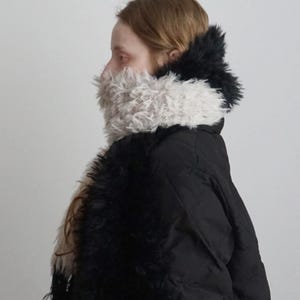 May include: A black quilted puffer jacket featuring a high collar and front panels adorned with long, shaggy faux fur in contrasting shades of cream and black. The textured fur trim extends from the neck down the front of the garment.