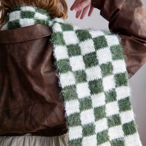 May include: A long, rectangular crochet scarf featuring a repeating checkered pattern in forest green and white. The scarf is constructed with a fuzzy, textured yarn and finished with long, white fringe tassels along the bottom edge.