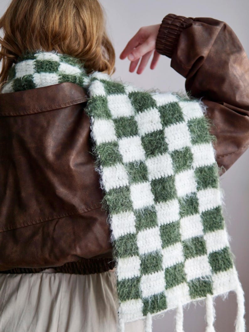 May include: A long, rectangular crochet scarf featuring a repeating checkered pattern in forest green and white. The scarf is constructed with a fuzzy, textured yarn and finished with long, white fringe tassels along the bottom edge.