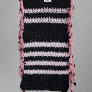 May include: A rectangular crochet scarf featuring alternating horizontal stripes of black and light pink yarn. The edges are adorned with long, dangling pink fringe accents. A small white rectangular label with the black text ZAIKA is attached.