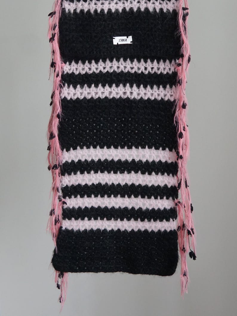 May include: A rectangular crochet scarf featuring alternating horizontal stripes of black and light pink yarn. The edges are adorned with long, dangling pink fringe accents. A small white rectangular label with the black text ZAIKA is attached.