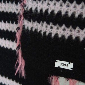 May include: A fuzzy knit garment featuring horizontal black and light pink stripes. The texture is soft and slightly brushed. A small white rectangular fabric label is stitched onto the lower right corner with the black text ZAIKA printed on it.