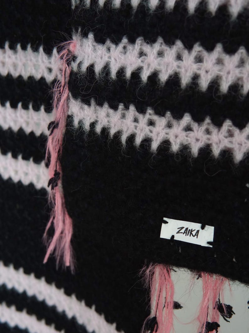 May include: A fuzzy knit garment featuring horizontal black and light pink stripes. The texture is soft and slightly brushed. A small white rectangular fabric label is stitched onto the lower right corner with the black text ZAIKA printed on it.