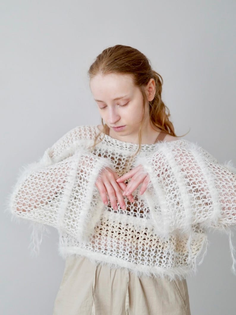 May include: A cream-coloured, loose-knit, open-weave jumper featuring long, wide sleeves with fuzzy, textured trim along the cuffs and hem. The garment has a wide neckline and a semi-sheer, airy construction with visible horizontal bands of varying knit density.
