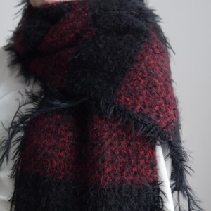 May include: A fuzzy, textured scarf featuring a bold, alternating block pattern of deep black and dark red. The material has a long-pile, shaggy appearance with loose, wispy fibres extending from the edges, creating a soft, voluminous look.