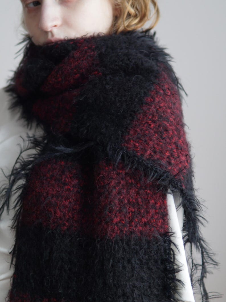 May include: A fuzzy, textured scarf featuring a bold, alternating block pattern of deep black and dark red. The material has a long-pile, shaggy appearance with loose, wispy fibres extending from the edges, creating a soft, voluminous look.